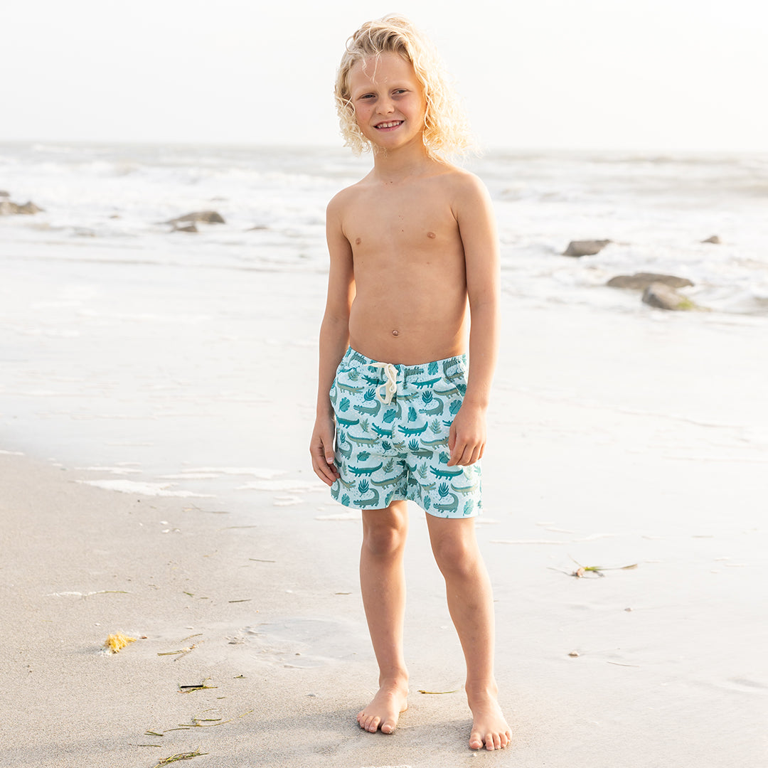 blonde boy smiling on the beach in his "later gator" swim trunks. the "later gator" print has a mix of light and dark green alligators, leaves, and white dots scattered on a teal colored background.