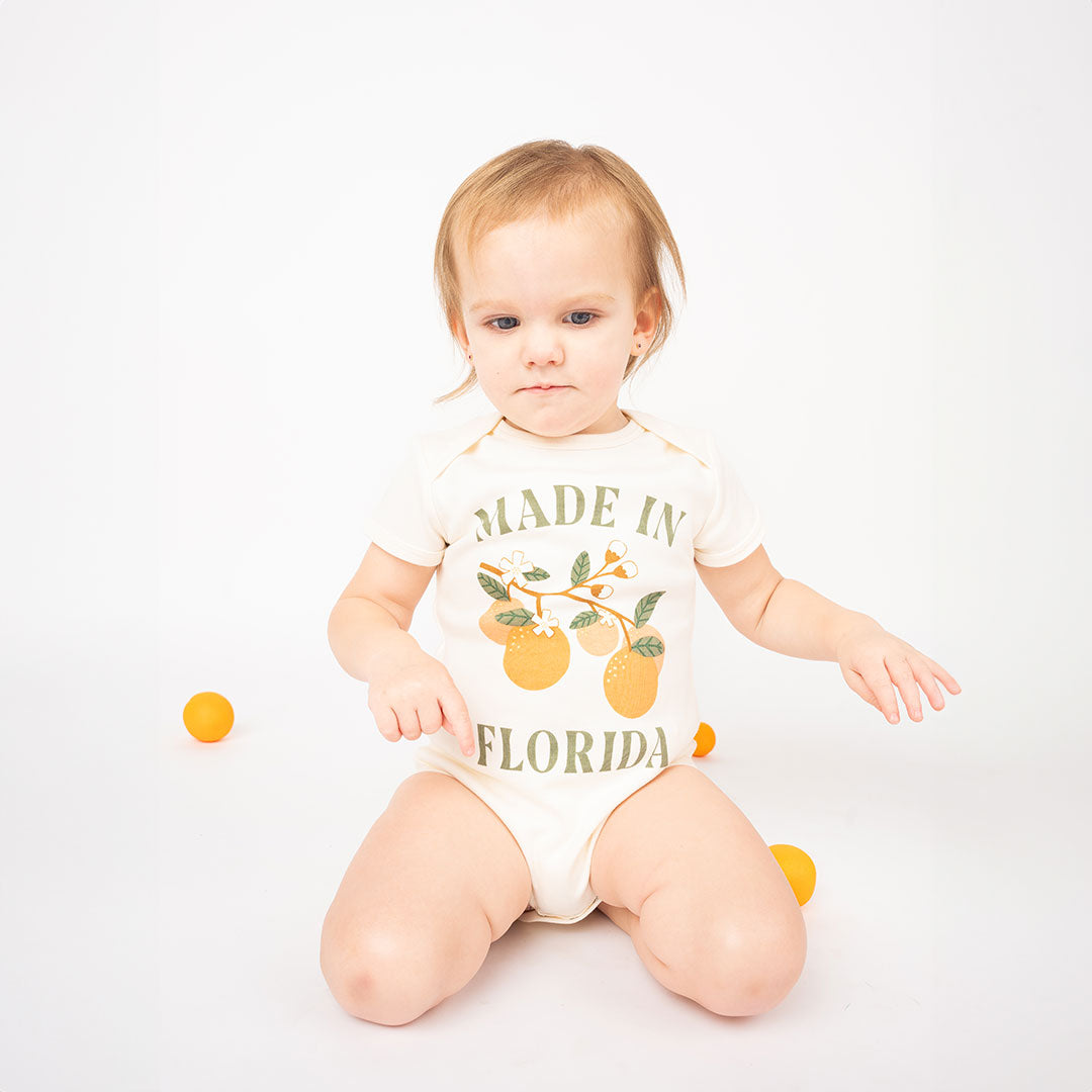 a baby sitting on their knees with oranges scattered around them. they are wearing the "made in Florida" cotton onesie. this cotton onesie has the phrase "made in Florida" in green print on the onesie. there is also a branch of oranges on it too.