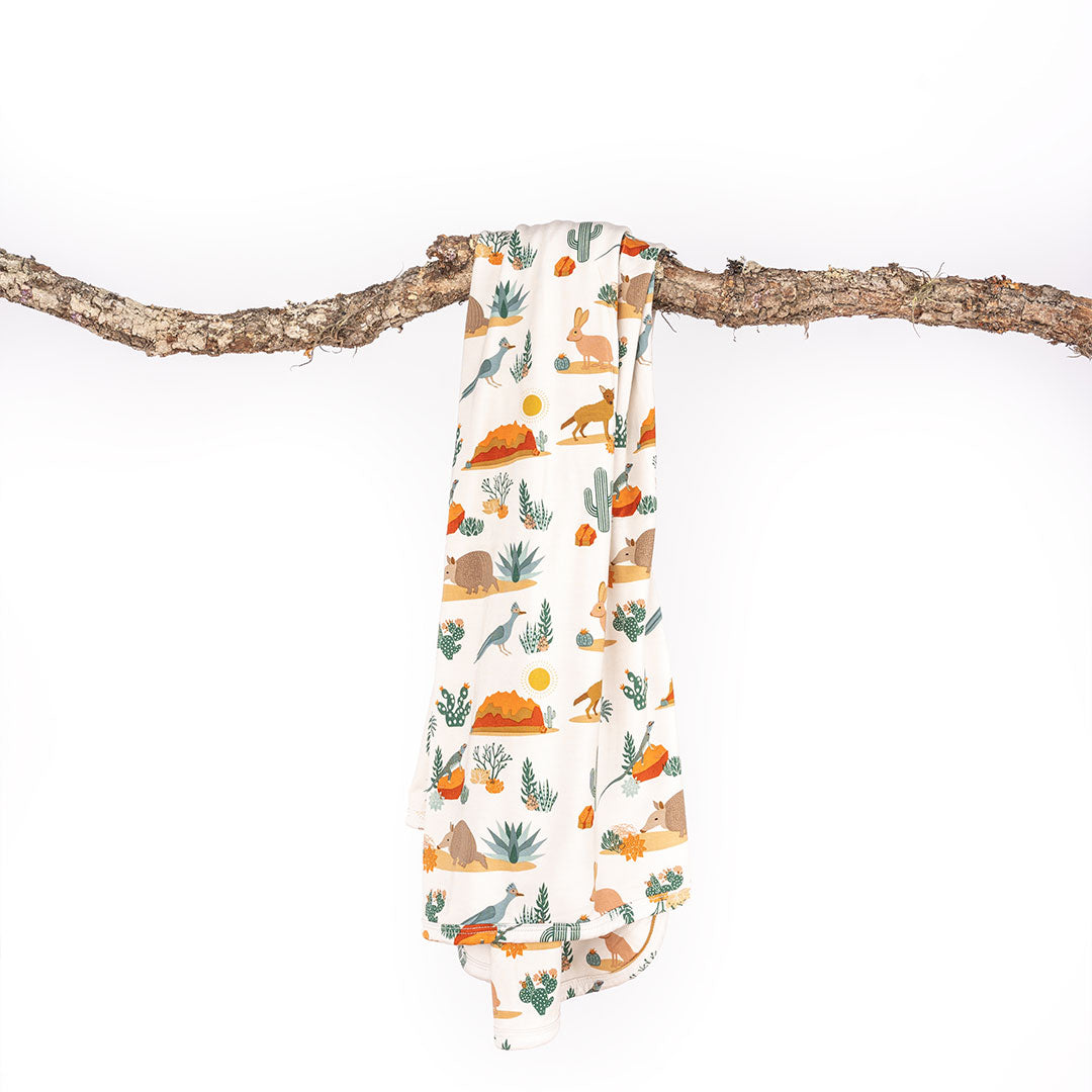 the "desert friends" bamboo blanket draped on a large tree stick. the "desert friends" print is a scattered pattern of armadillo, cacti, other desert plants, rocks, desert foxes, lizards, and desert birds. this is all on a beige background.