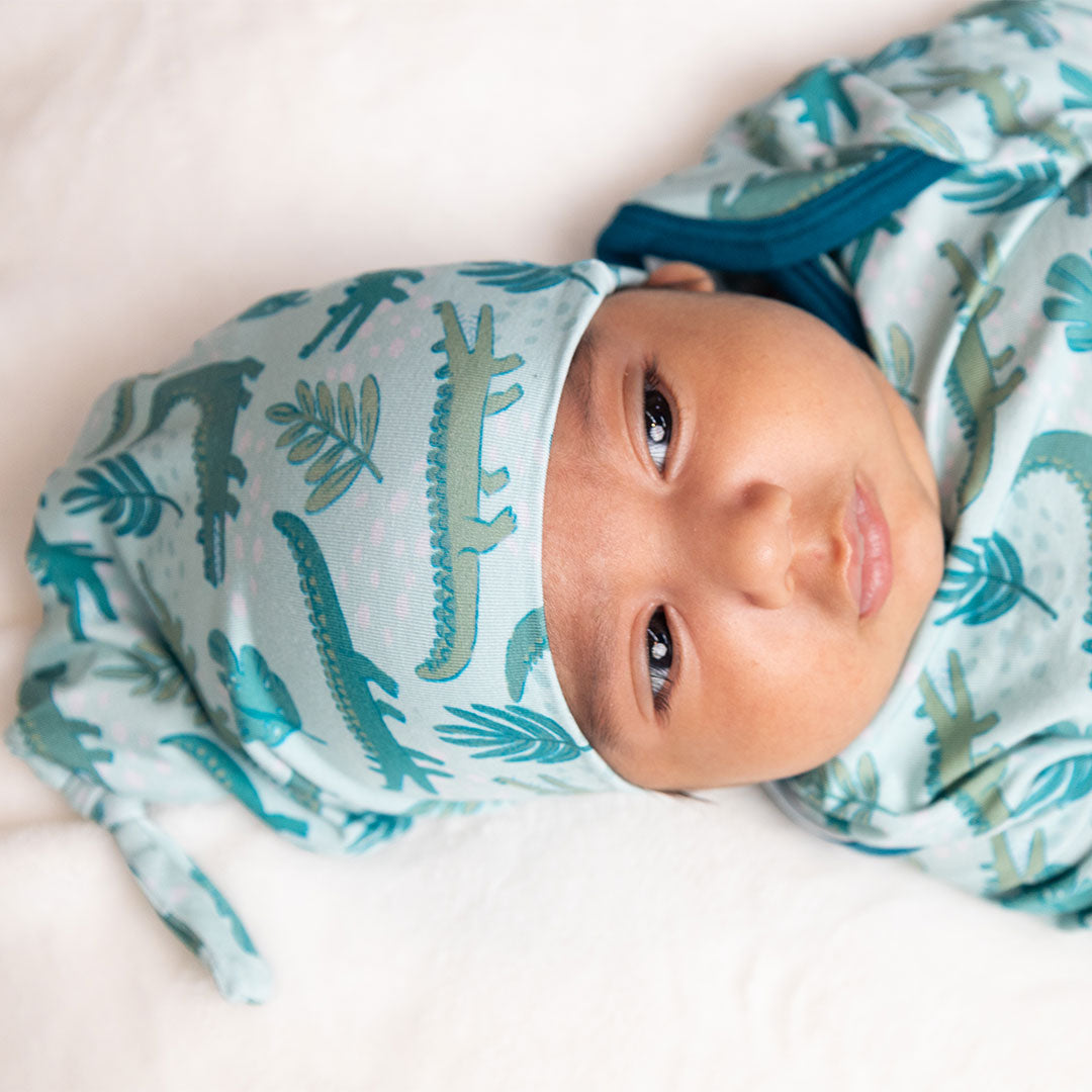 little baby in his "later gator" hat. the "later gator" print has a mix of light and dark green alligators, leaves, and white dots scattered on a teal colored background.