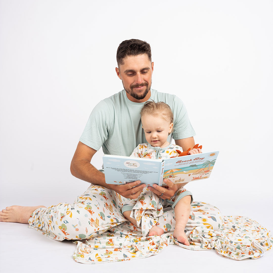 a father reads the book "beach day" to his baby son. he wears the "beach days" relaxed pants and the baby wears the matching shortie romper. they are sitting on a the "beach days" blanket. the "beach days" print is a scattered print of beach umbrellas, beach balls, sand buckets and shovels, coral, shells, beach chairs, seagulls, flipflops, and sunglass mixed in a orderly and creative way.