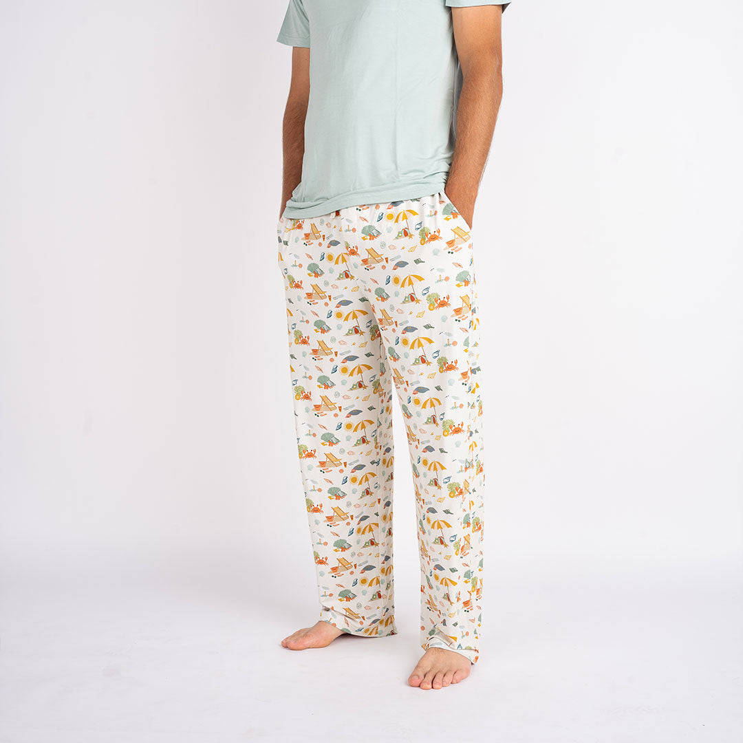 a man posing in the "beach days" relaxed pants. his hands are in the pockets of the pants. the "beach days" print is a scattered print of beach umbrellas, beach balls, sand buckets and shovels, coral, shells, beach chairs, seagulls, flipflops, and sunglass mixed in a orderly and creative way.