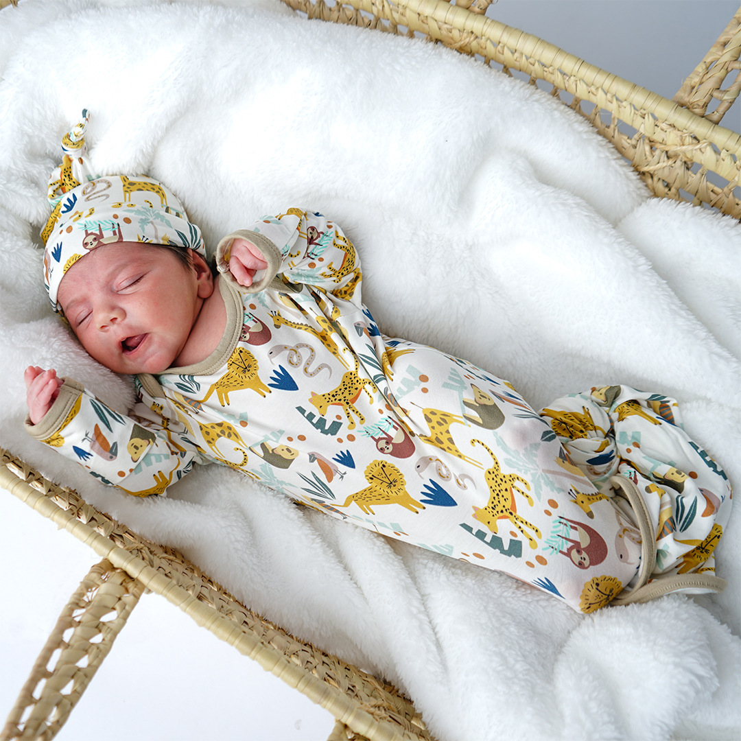 a baby lays in a basket cradle, on a white blanket. the baby wears the "jungle friends" matching gown and hat set. the "jungle friends" print is a mix of jungle animals that include giraffes, lions, cheetahs, sloths, snakes, and forest plants.