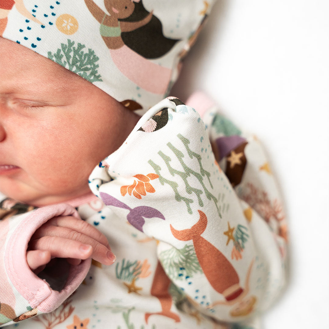 a close up and focused photo of half a babies upper body. you can see their fingers, and head. the infant is wearing the "making waves" gown and hat set. the "making waves" print has a diverse spread of mermaids, sea coral, starfish, fish, and bubbles all spread out in different colors. this is all put on a beige background.