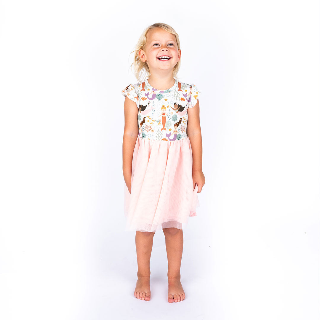 a young girl stands laughing in her "making waves" tulle twirl dress. the "making waves" print has a diverse spread of mermaids, sea coral, starfish, fish, and bubbles all spread out in different colors. this is all put on a beige background.