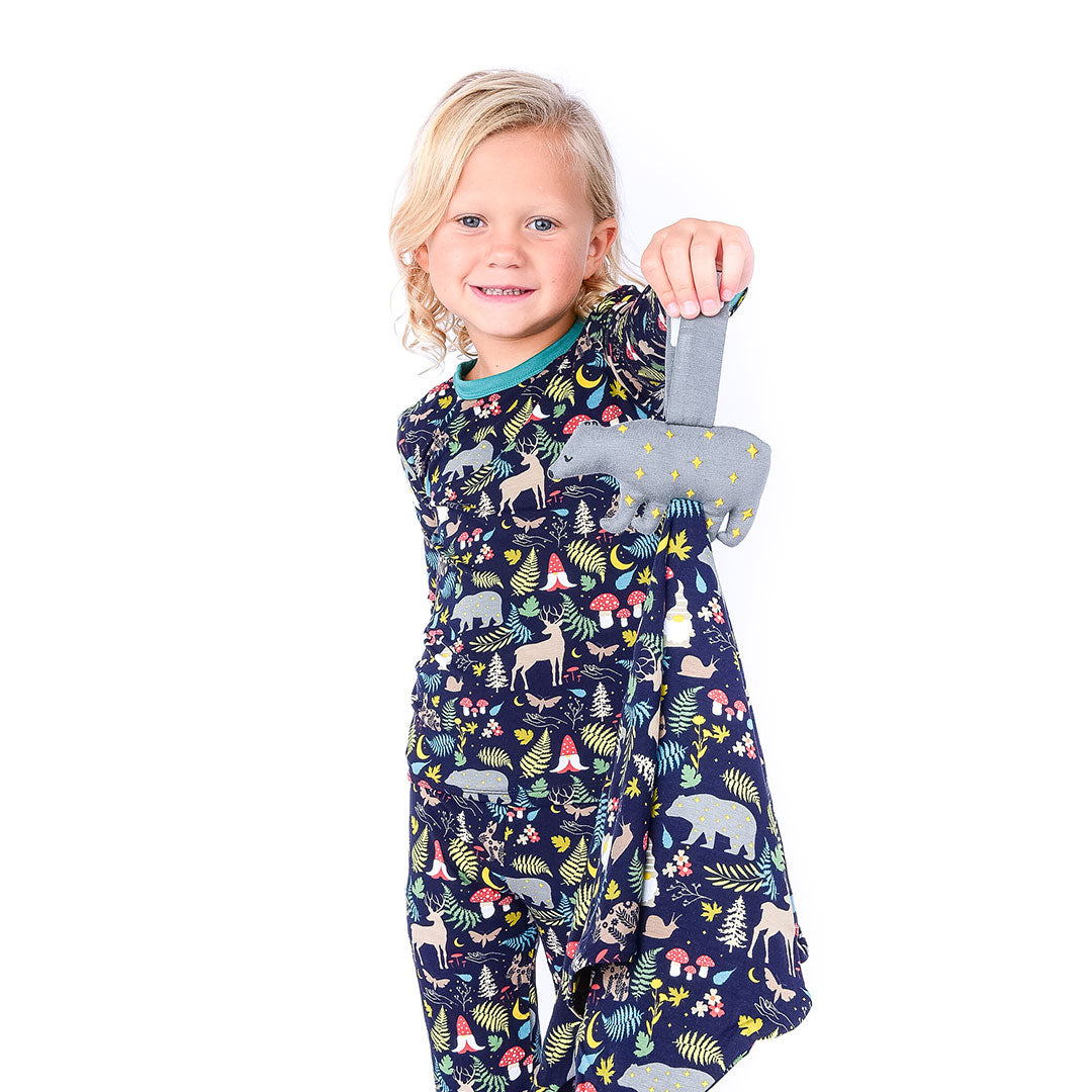 little girl holds out the "night forest" lovey. she is wearing the 2-piece matching set. the "night forest" print is a night time themed design. you can see an array of forest animals ranging from, deer, bears, bunnies, birds, forest trees and leaves, flowers, and mushrooms. there are also starts and moons scattered around to enhance the night time atmosphere.