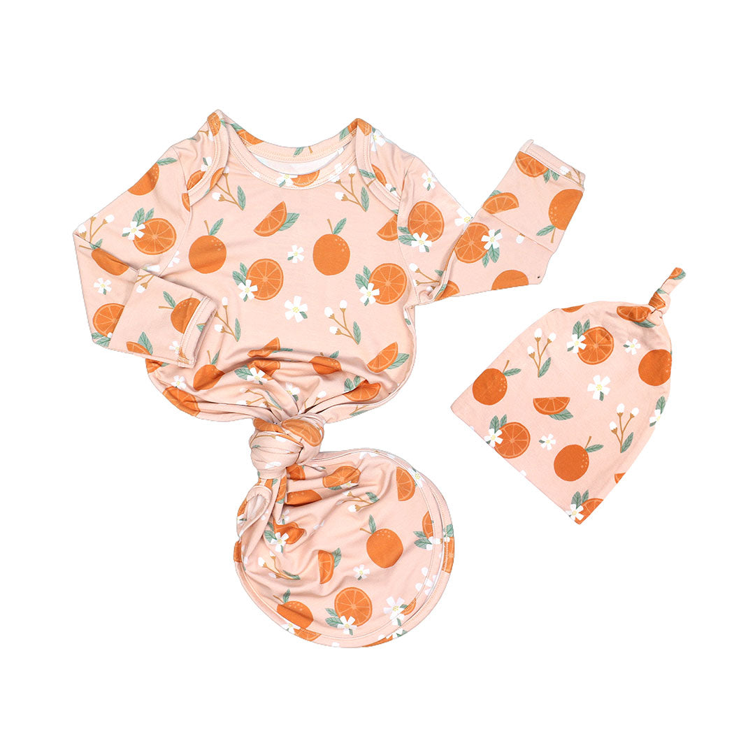 the "freshly squeezed" bamboo gown and hat newborn set. the bottom half of the gown is tied in a knot. the "freshly squeezed" print has an assortment of full and half cut oranges scattered around. there is also flower heads and flower stems that intermingle within the print. this is all space out around a pink background space.