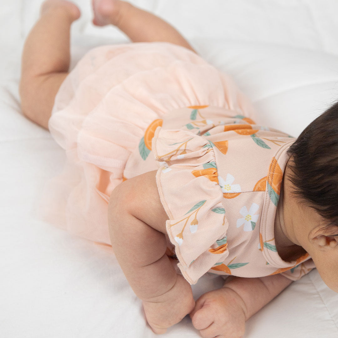 a zoomed in photo of a baby play on its tummy, wearing the "freshly squeezed" skirted onesie. this picture highlights the fact that the skirt wraps all the way around the onesie. the "freshly squeezed" print has an assortment of full and half cut oranges scattered around. there is also flower heads and flower stems that intermingle within the print. this is all space out around a pink background space.