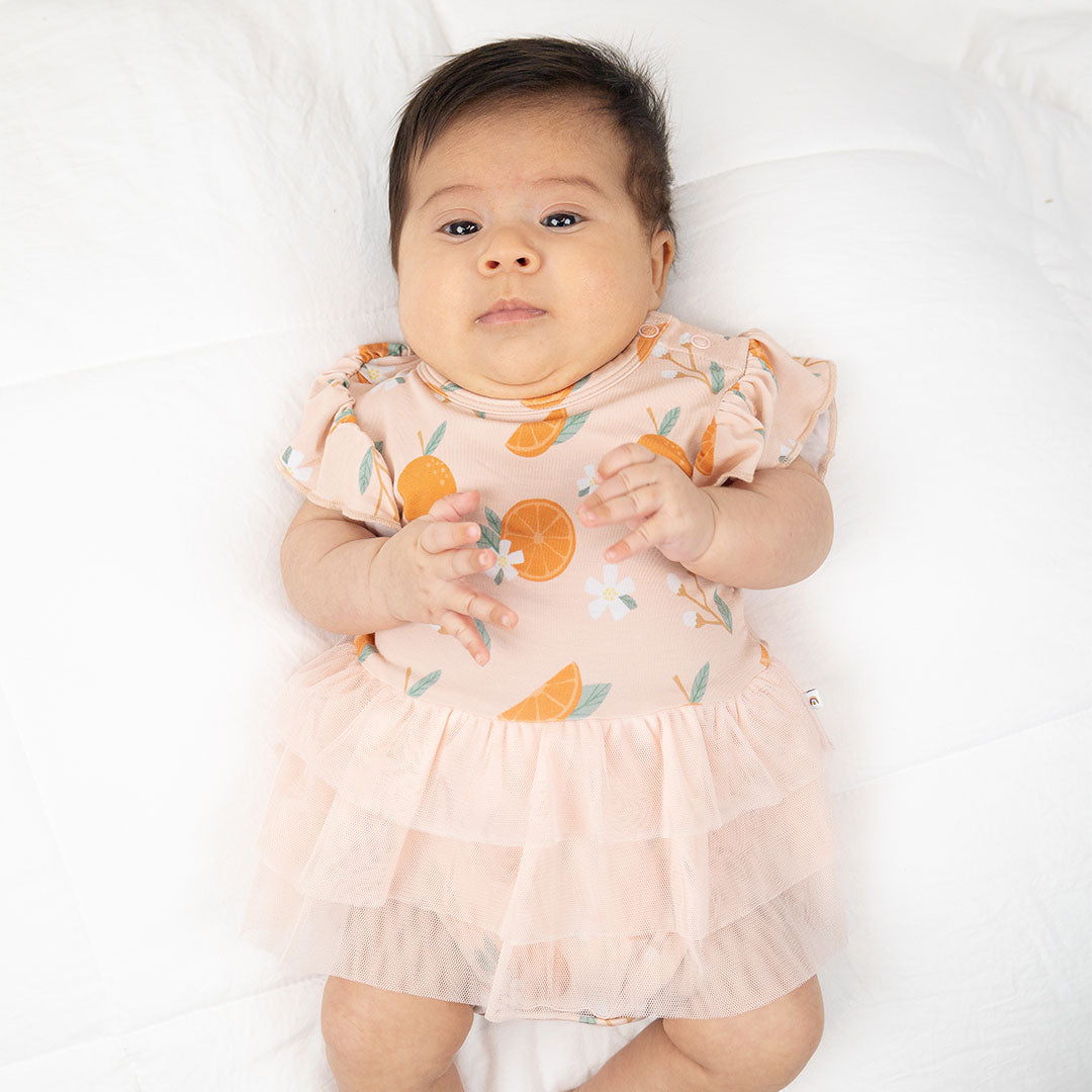 a baby is laying on its back in the "freshly squeezed" skirted onesie. the "freshly squeezed" print has an assortment of full and half cut oranges scattered around. there is also flower heads and flower stems that intermingle within the print. this is all space out around a pink background space.