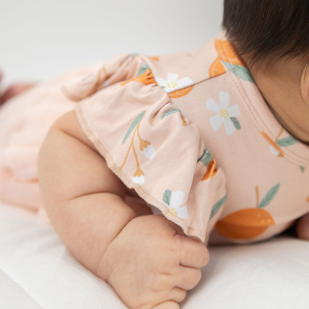 zoomed in photo of a babies fist and shoulder in the "freshly squeezed" skirted onesie. the "freshly squeezed" print has an assortment of full and half cut oranges scattered around. there is also flower heads and flower stems that intermingle within the print. this is all space out around a pink background space.
