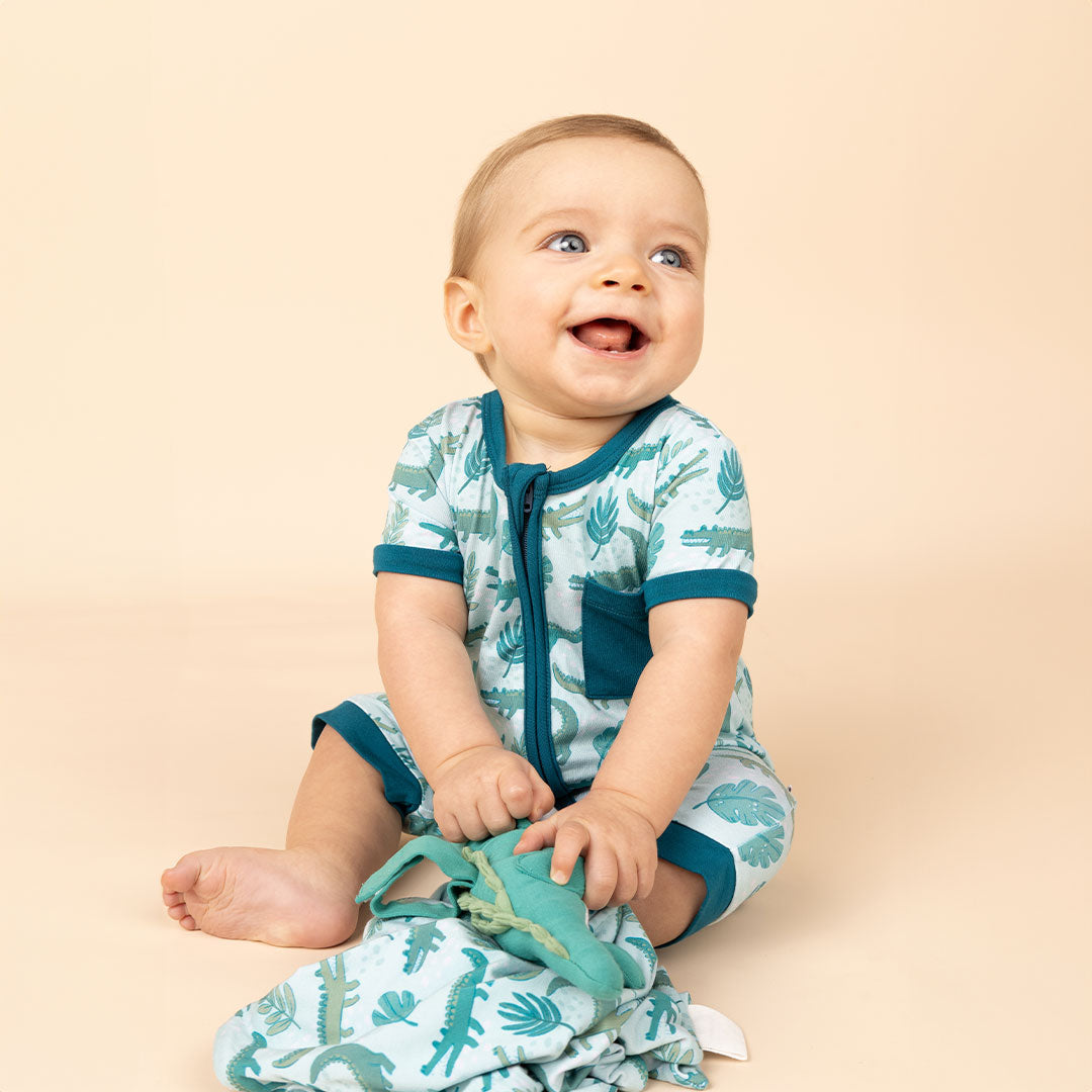 a happy baby in his "later gator" shortie. he is playing with his "later gator" lovey. the "later gator" print has a mix of light and dark green alligators, leaves, and white dots scattered on a teal colored background.