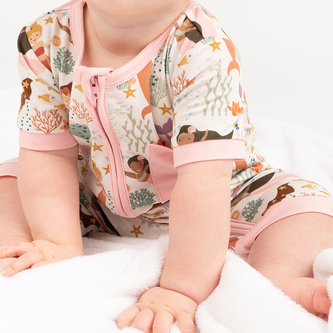 an up close, focused picture of a baby sitting on the floor. she is wearing the "making waves" shortie. the "making waves" print has a diverse spread of mermaids, sea coral, starfish, fish, and bubbles all spread out in different colors. this is all put on a beige background.