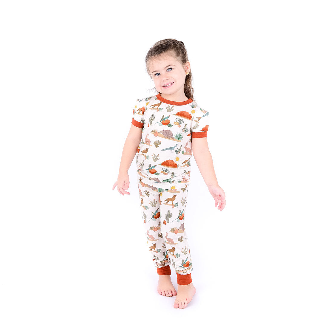 toddler girl poses in her "desert friends" 2-piece matching pajama set. the "desert friends" print is a scattered pattern of armadillo, cacti, other desert plants, rocks, desert foxes, lizards, and desert birds. this is all on a beige background.