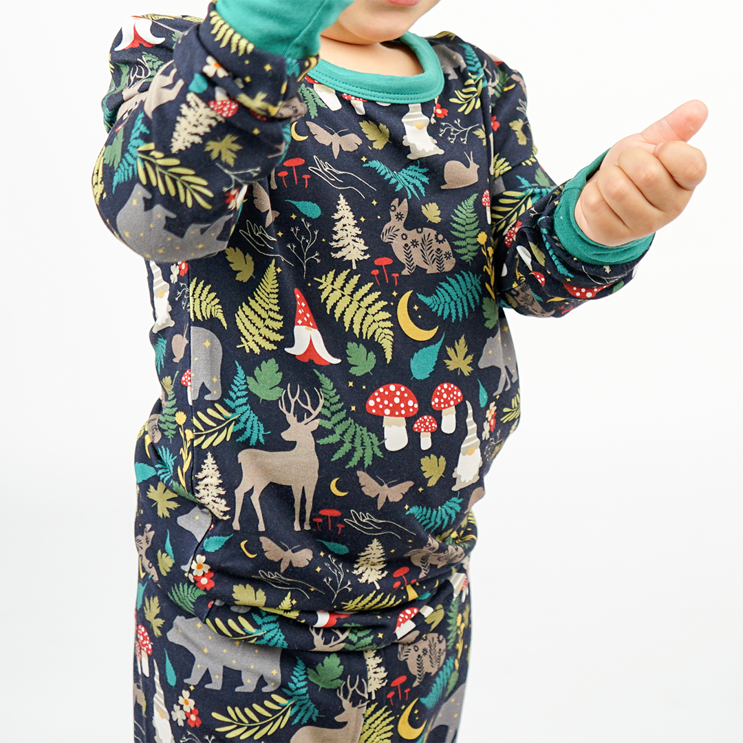 a close up picture of a child wearing the "night forest" 2-piece matching pajama set. the "night forest" print is a night time themed design. you can see an array of forest animals ranging from, deer, bears, bunnies, birds, forest trees and leaves, flowers, and mushrooms. there are also starts and moons scattered around to enhance the night time atmosphere.