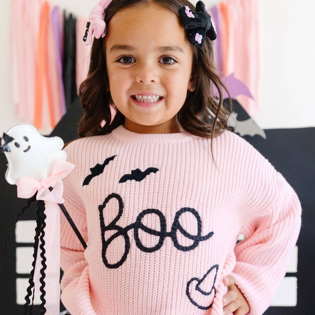 little girl wearing a 2 pack of Halloween themed hair clips. One is velvet black material with sequins stitched onto the ends and the word BOO in pink letters, and the other clip is pink velver with sequins and the word boo in black letters