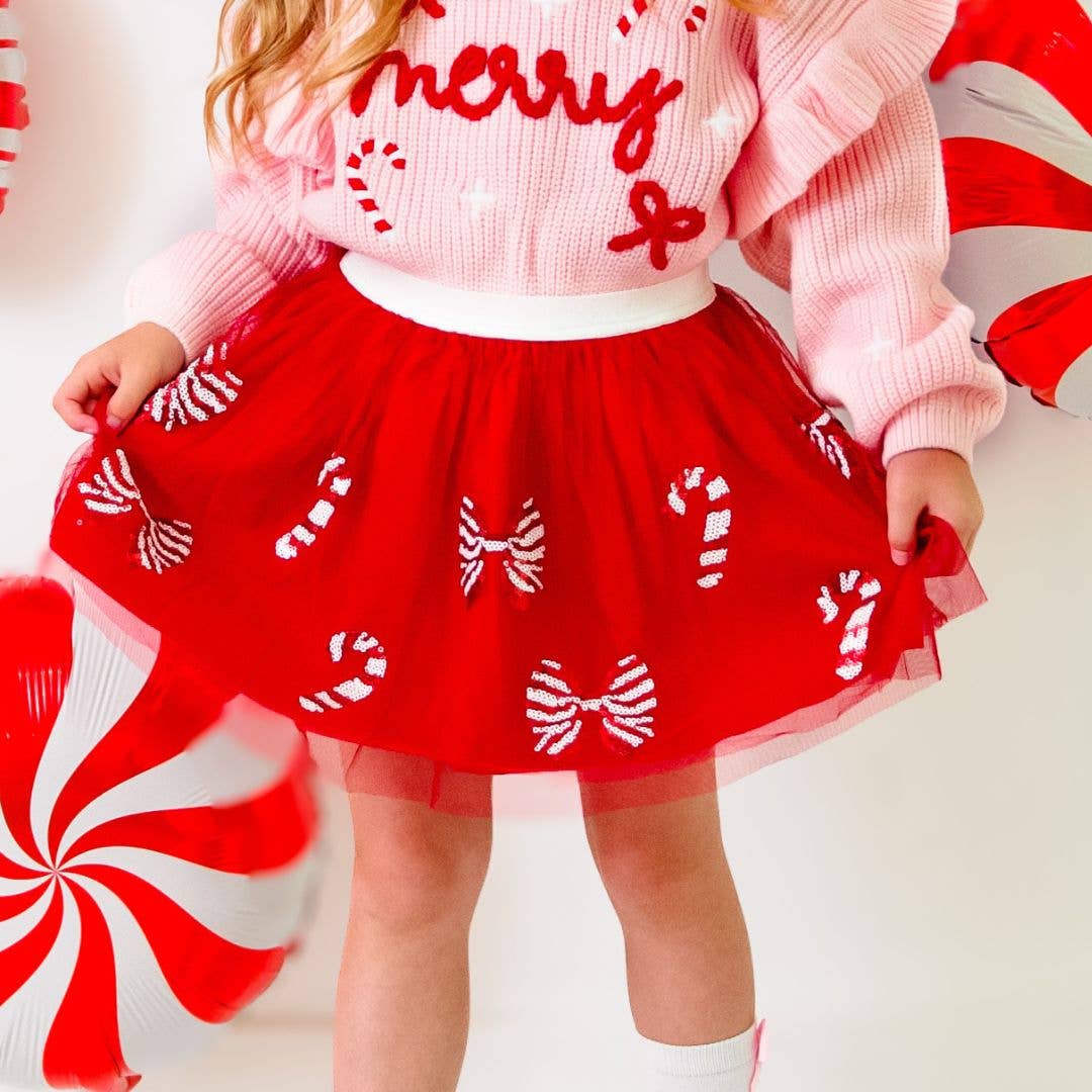Child wearing a festive outfit with a pink sweater that says “merry” and a red tulle tutu skirt decorated with sequin candy canes and striped bows, paired with white socks.