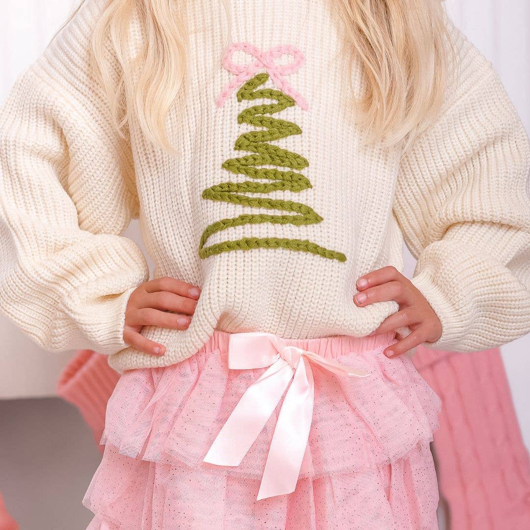 Child wearing a cream knit sweater with a green Christmas tree design topped with a pink bow, paired with a layered pink tulle skirt featuring a satin ribbon bow at the waist.