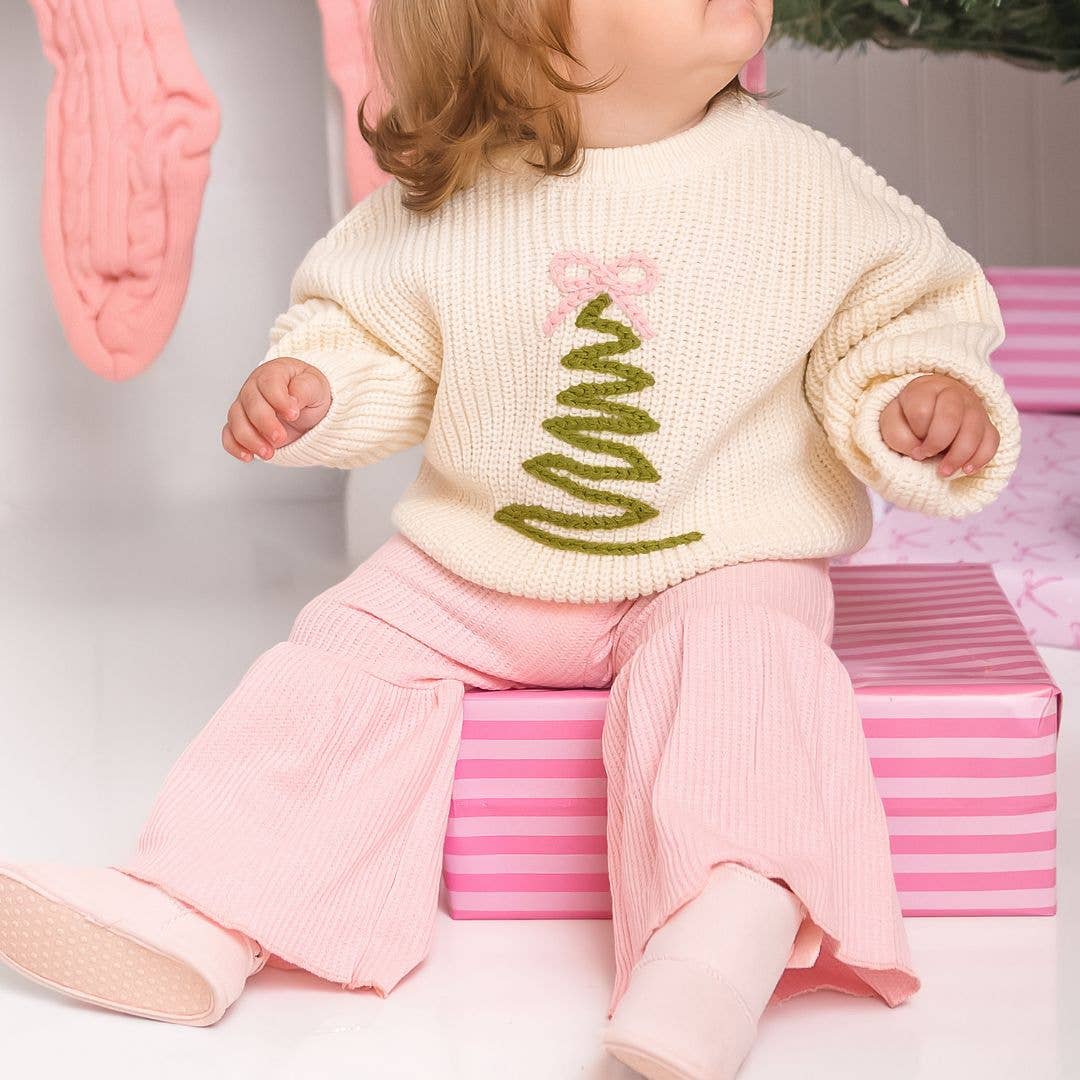 Baby wearing a cream knit sweater with a green Christmas tree design topped with a pink bow, paired with light pink ribbed pants and soft pink shoes, sitting on a pink striped gift box.
