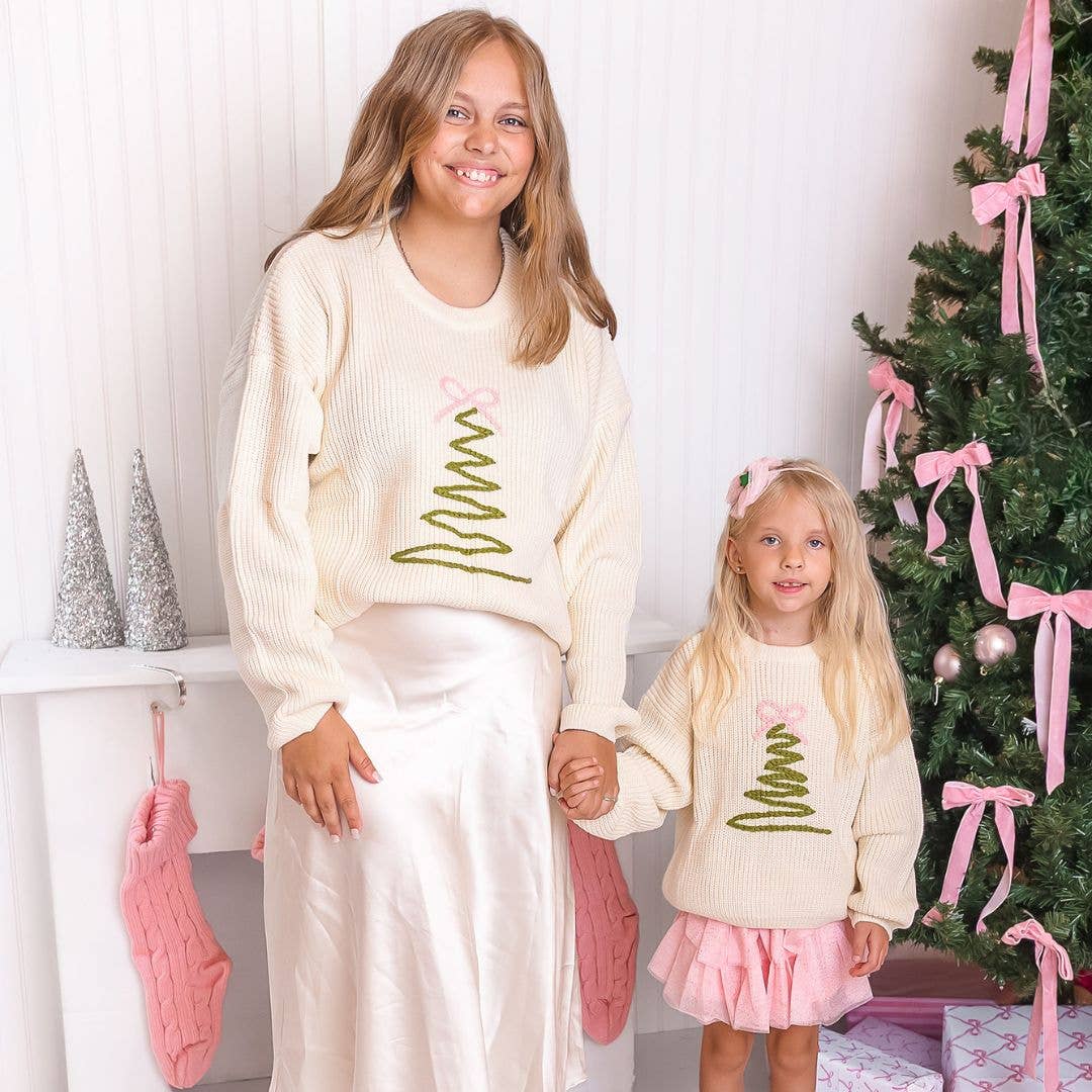 Woman and young girl standing together in matching cream-colored knit sweaters featuring a simple green zigzag Christmas tree topped with a pink bow. The woman wears a long ivory satin skirt, while the girl wears a pink skirt with a bow headband. They are standing by a Christmas tree decorated with pink bows and ornaments, with pink stockings and wrapped presents nearby.