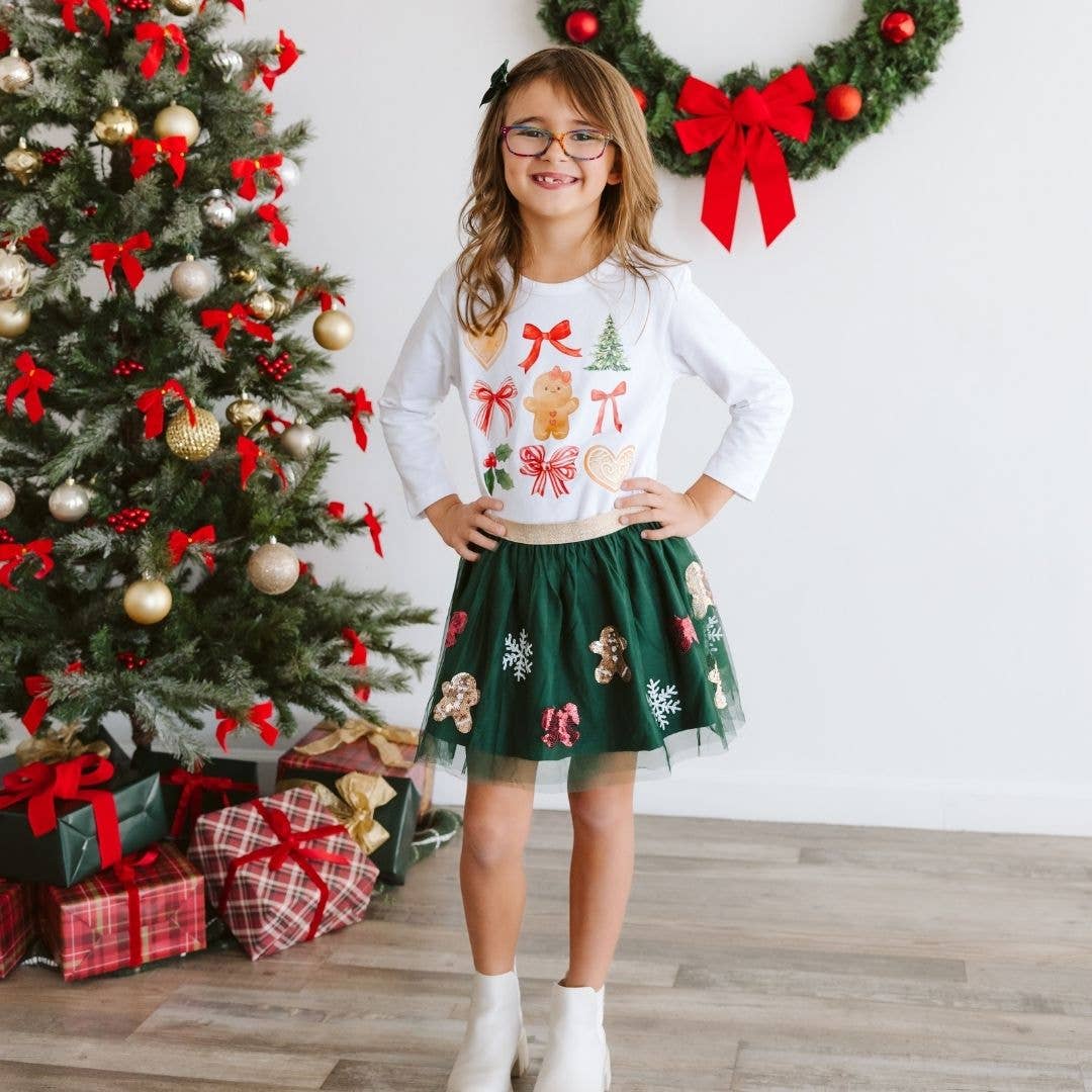 Smiling child wearing a festive outfit with a white long-sleeve Christmas shirt decorated with bows, a gingerbread girl, holly, and cookies, paired with a green tulle skirt featuring gingerbread men, candy canes, and snowflakes, standing in front of a decorated Christmas tree and wrapped presents.