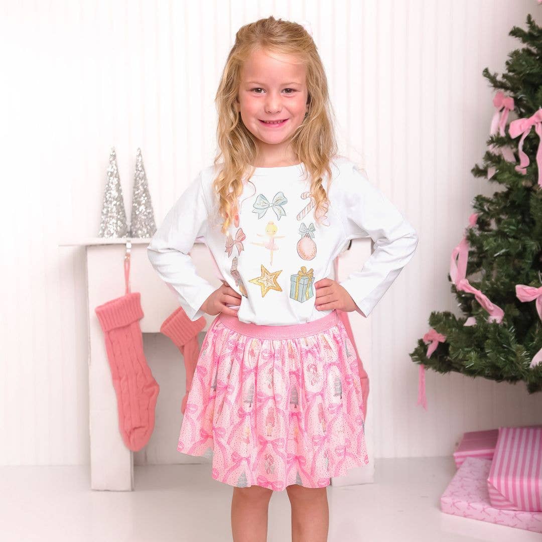 Young girl standing indoors wearing a festive outfit with a white long-sleeve shirt decorated with pastel holiday icons such as bows, candy canes, ornaments, and gifts, paired with a pink tutu skirt patterned with nutcrackers, Christmas trees, and bows. She is smiling with hands on her hips, standing near a Christmas tree decorated with pink ribbons, silver cone decorations, and pink knit stockings.