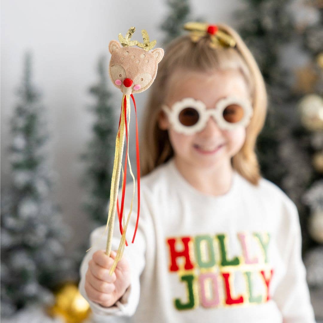 Child holding a reindeer-themed Christmas wand with gold antlers, a red pom-pom nose, and red and gold ribbon streamers, while wearing festive sunglasses and a white sweater with the words “HOLLY JOLLY” in red and green letters.