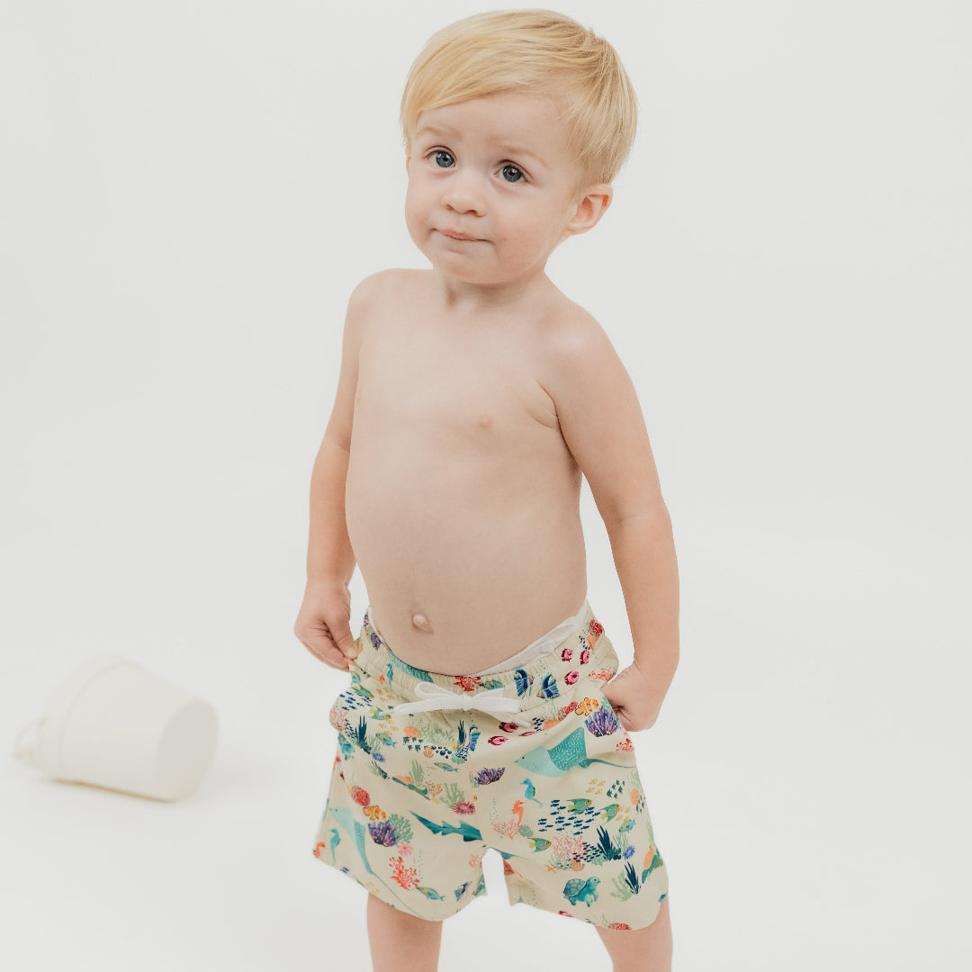 Toddler boy wearing our Emerson and Friends Coral Reef Boys Swim Trunks, featuring a vibrant ocean-themed design with sharks, sea turtles, and coral, made from quick-dry fabric with UPF sun protection for all-day beach and pool fun.