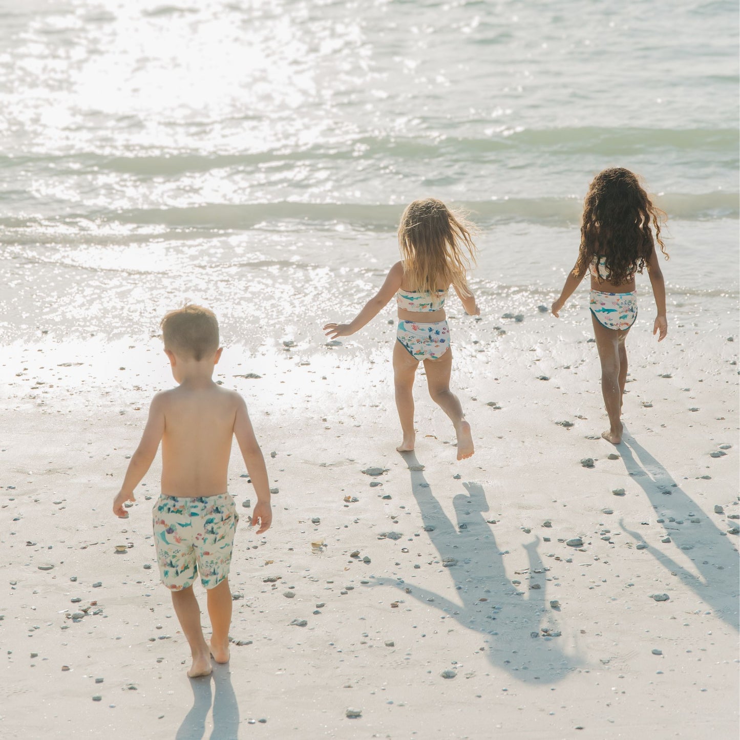 Toddler friends walking into the beach in a Coral Reef Bikini Swimsuit featuring a vibrant two-piece ocean print with sharks and sea turtles, made from breathable, UPF 50+ fabric for sunny beach adventures.