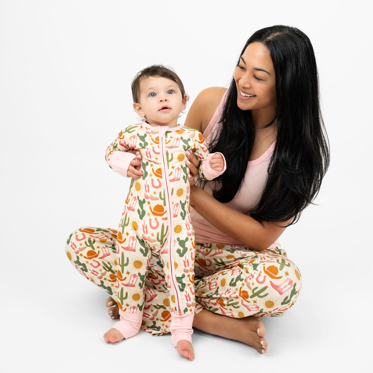 Baby girl and mom wearing our Emerson and Friends Hey Y’all Convertible Bamboo Convertible Pajamas and Hey Y'all relaxed pants. The print shows green cactus, yellow sun, brown cowgirl hat, pink boots and horseshoes. Perfect for cozy rodeo nights or pajama parties with a cowgirl flair!
