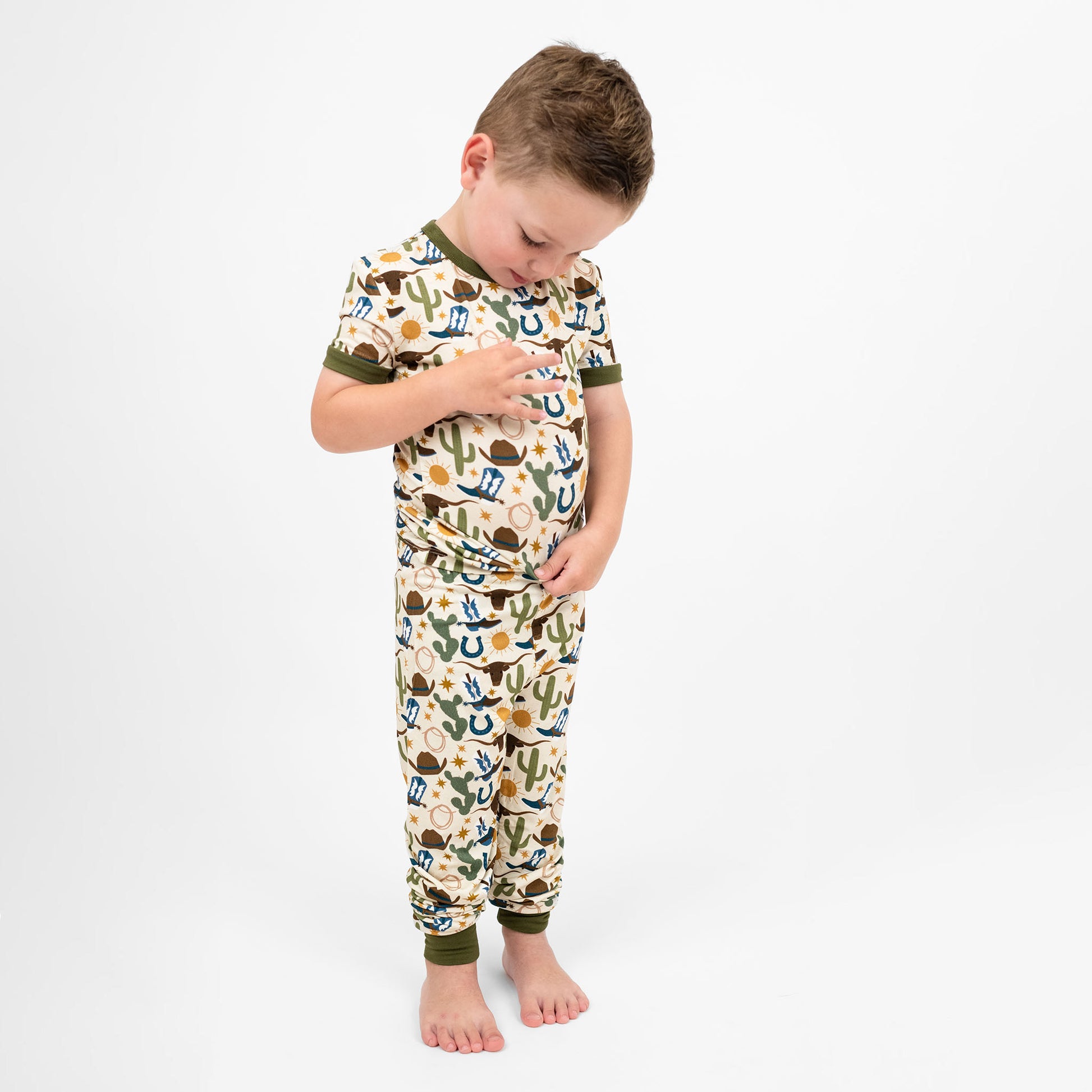 Toddler boy pointing our Western Howdy Collection! This cozy bamboo 2-piece pajama features a charming cowboy-themed print with blue boots, cacti, suns, horseshoes, and cowboy hats. Perfect for western cozy vibes.