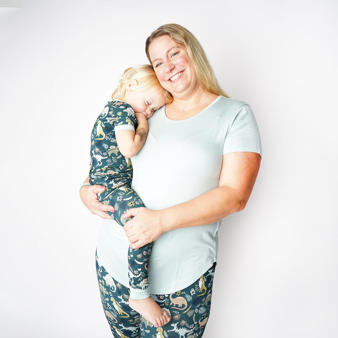 Mom and daughter pose. the mom holds her baby while the baby lays her head on her moms shoulder. the mom is wearing the "blue surf" women's top and the "dino" joggers. the baby is wearing the "dino" 2-piece matching pajama set.