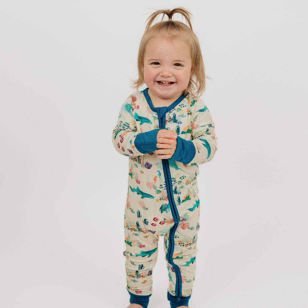 Baby girl wearing our Coral Reef Bamboo Convertible Baby Pajamas by Emerson and Friends, featuring a vibrant ocean-themed print with sharks, sea turtles, and coral reefs. Made from ultra-soft bamboo fabric with fold-over cuffs and a two-way zipper for easy diaper changes.