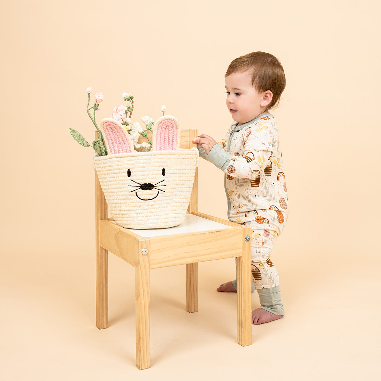 Baby standing, wearing Hop to It convertible pajama with easter print, touching white easter basket.