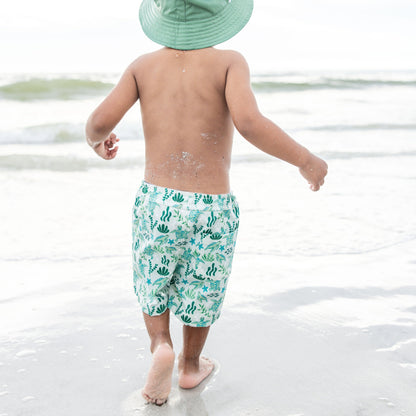 Boy waking on the beach wearing our Save the Sea Turtle Swim trunks. The print is featuring a sea-green turtle and ocean-inspired print. Made from quick-dry, lightweight fabric with an adjustable drawstring waist for a comfy fit. Perfect for beach days and poolside fun!