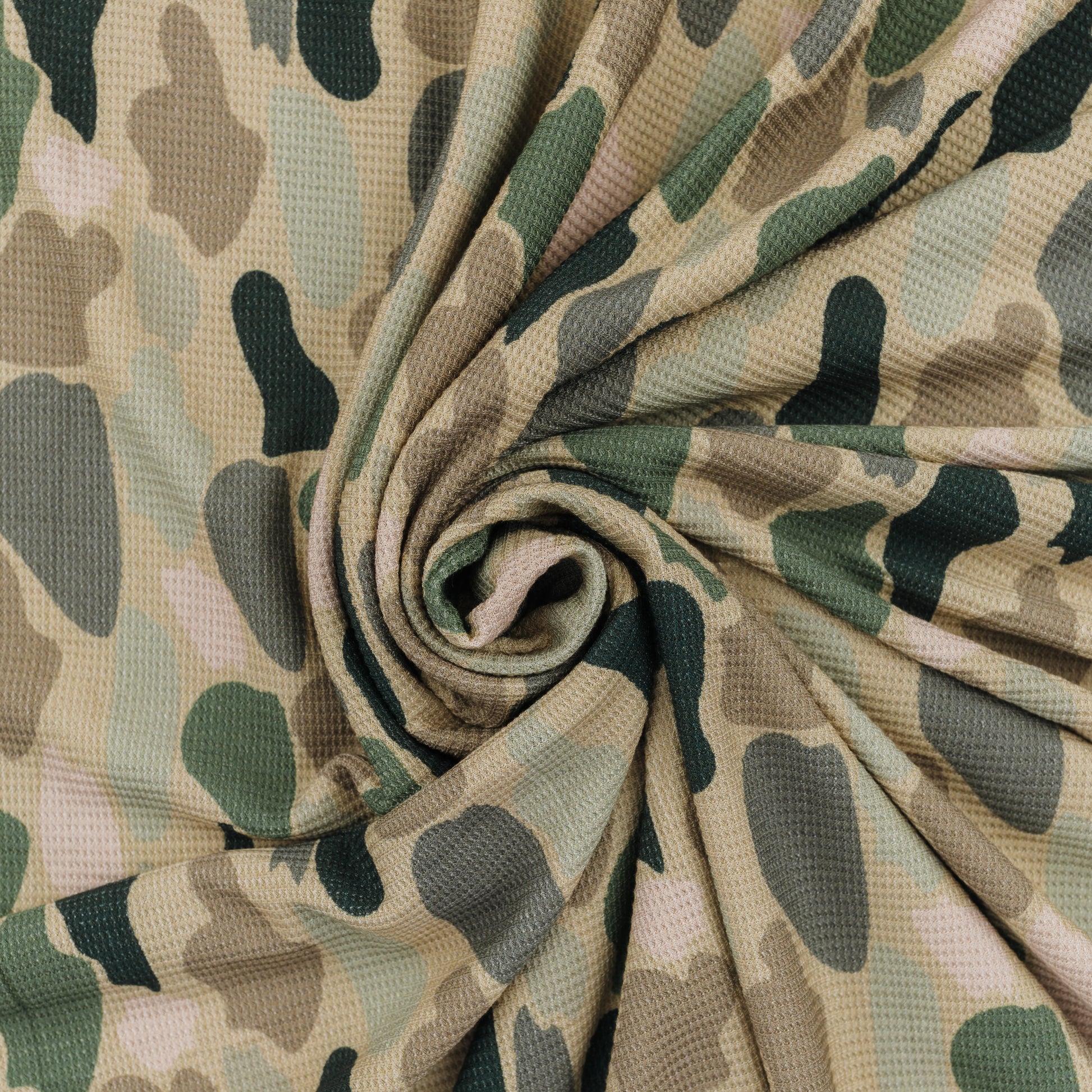 A close-up of camouflage-patterned blanket arranged in a spiral drape, showing a textured waffle-weave surface with shades of beige, green, and gray.