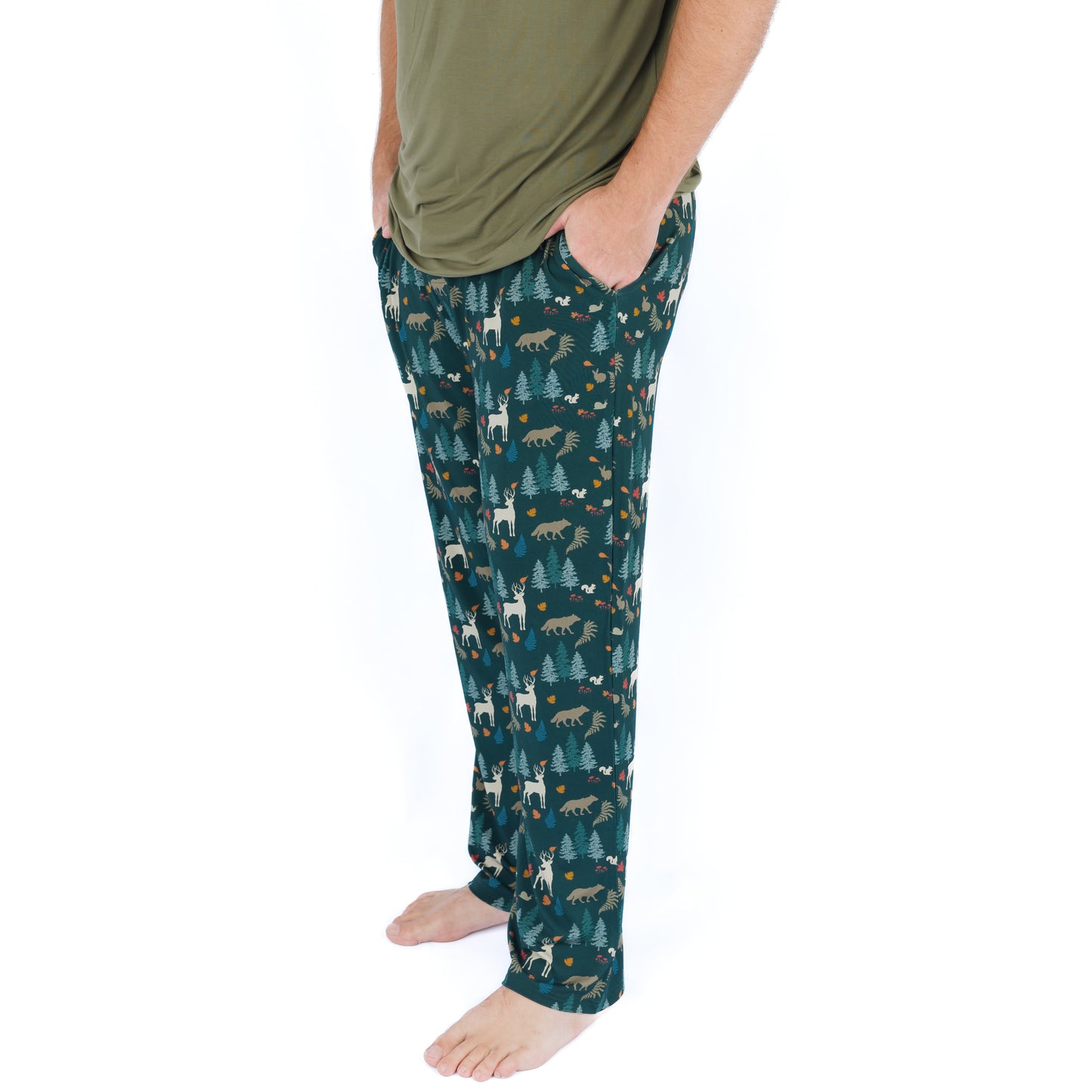A man wearing dark teal patterned pajama pants with a forest print and an olive green t-shirt, standing against a white background.