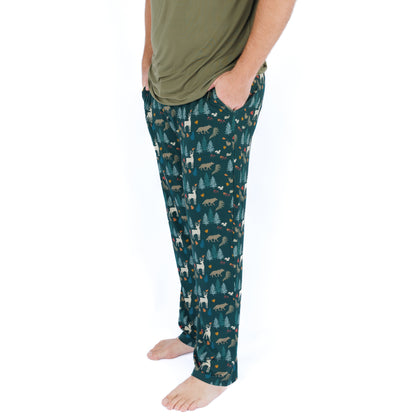 A man wearing dark teal patterned pajama pants with a forest print and an olive green t-shirt, standing against a white background.