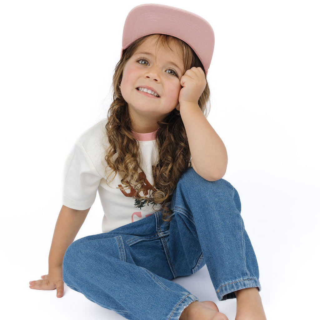 A young girl with long curly brown hair sits barefoot on a white studio background, wearing a pink baseball cap, a white graphic T-shirt, and blue jeans.