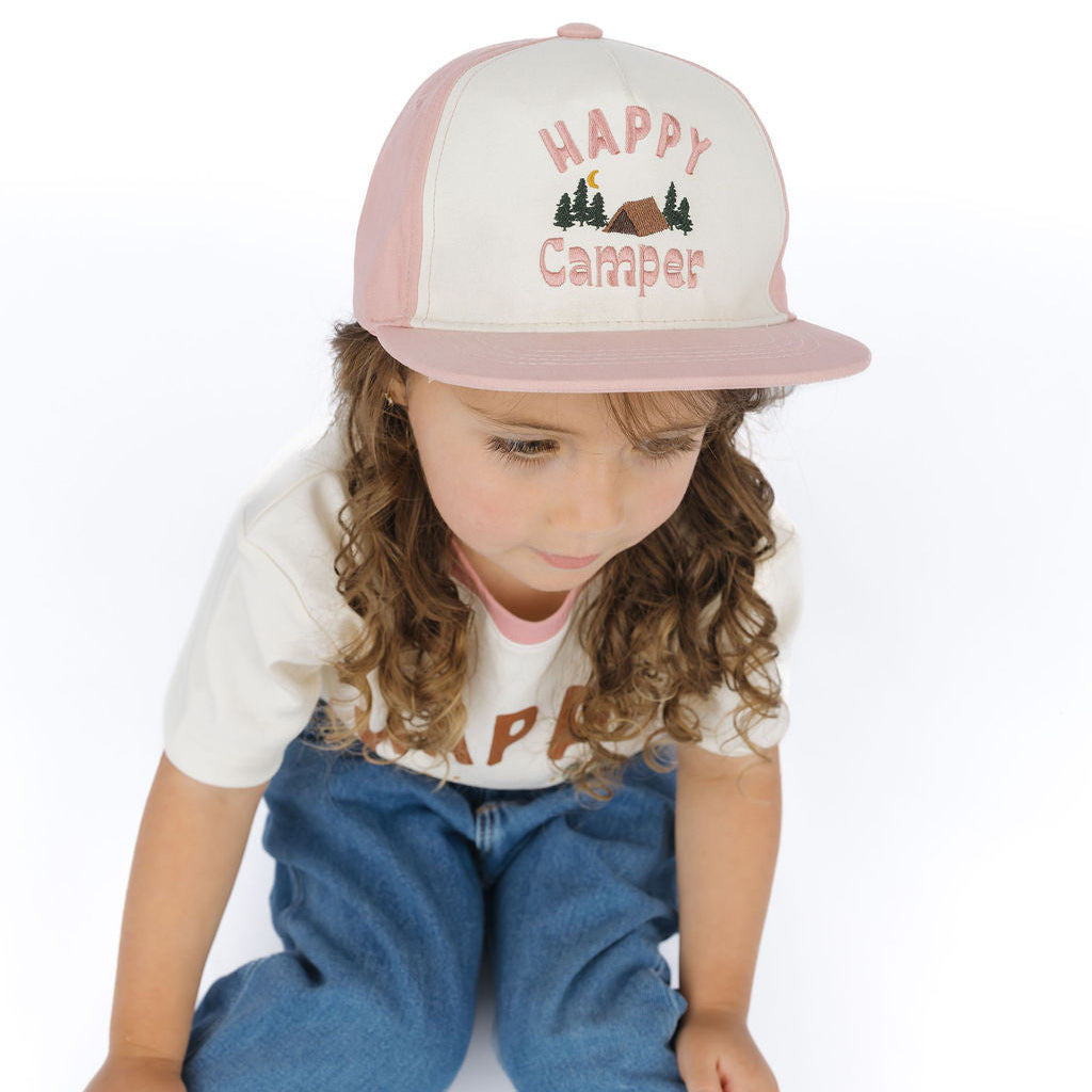 A young child wearing a pink and white trucker-style cap with 'HAPPY Camper' embroidered on the front, sitting against a plain white background.