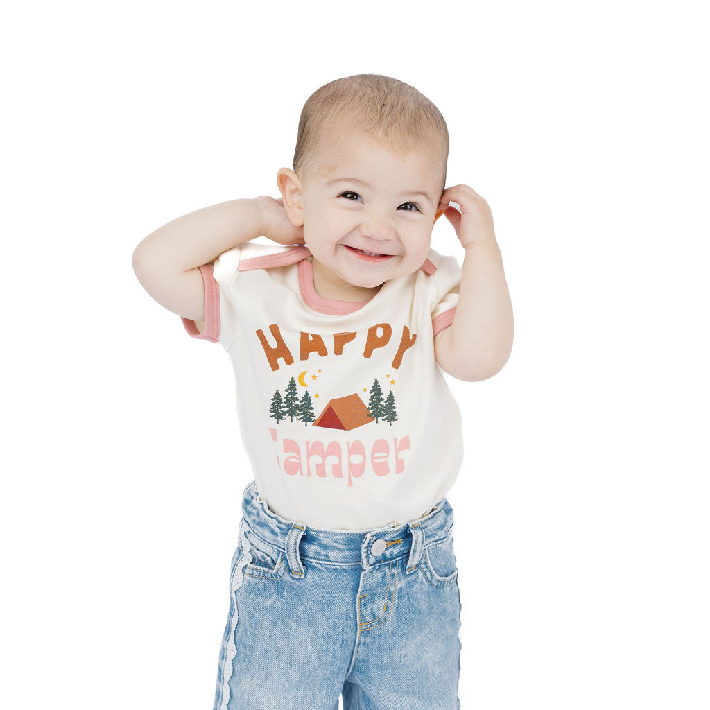 A smiling baby standing against a white background, wearing a cream-colored short-sleeve t-shirt with a colorful 'Happy Camper' graphic and light blue denim pants.