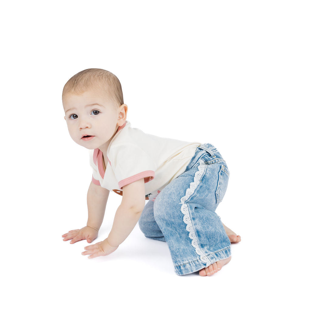 A baby crawling on a white studio background, wearing a cream ringer t-shirt with pink trim and light blue denim jeans with white lace trim down the leg.