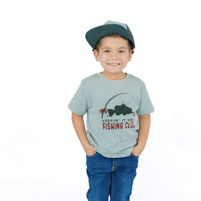 A young boy standing against a white background, wearing a light blue graphic t-shirt with a fishing design and the text 'KEEPIN' IT FOR FISHING CLUB.', blue jeans, and a dark green cap.