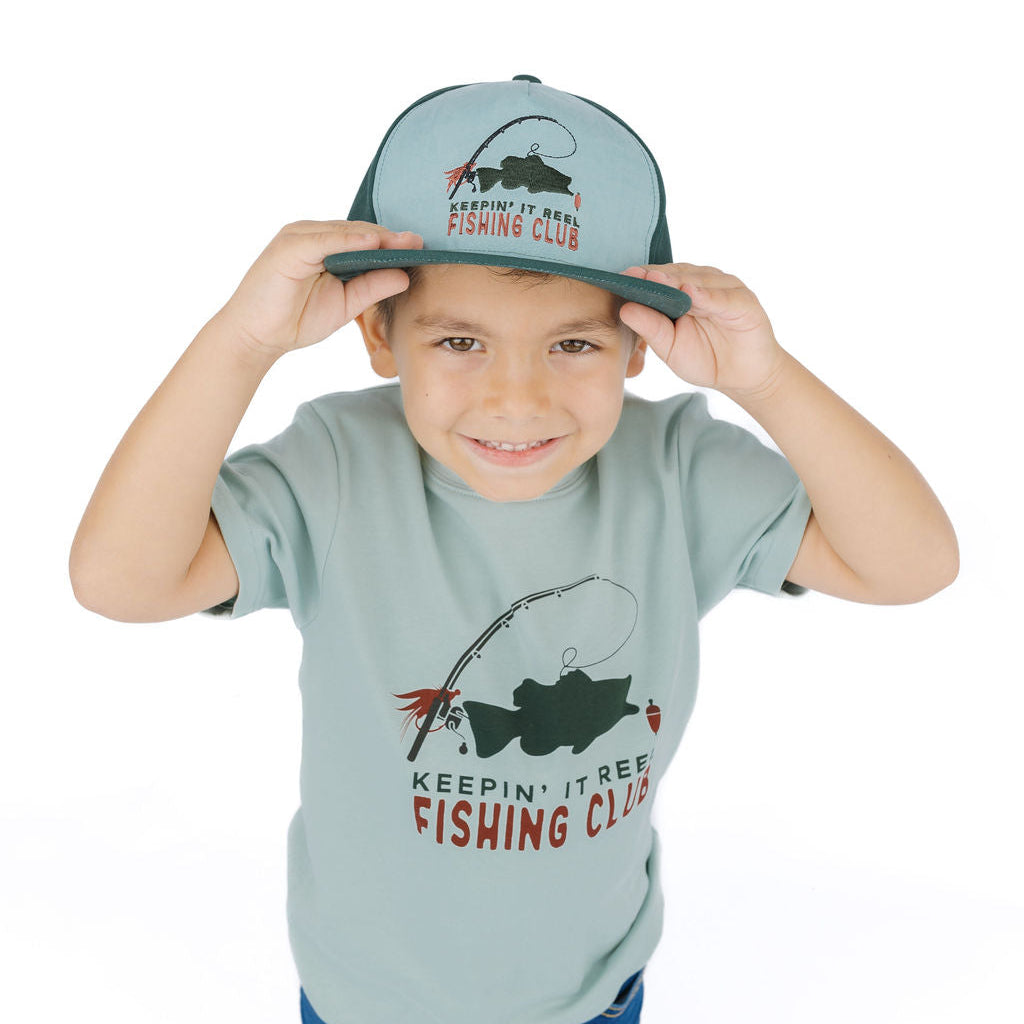 A young boy wearing a light sage green t-shirt and matching cap with a fishing graphic and the text 'KEEPIN' IT BEE FISHING CLUB'.