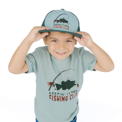 A young boy wearing a light sage green t-shirt and matching cap with a fishing graphic and the text 'KEEPIN' IT BEE FISHING CLUB'.