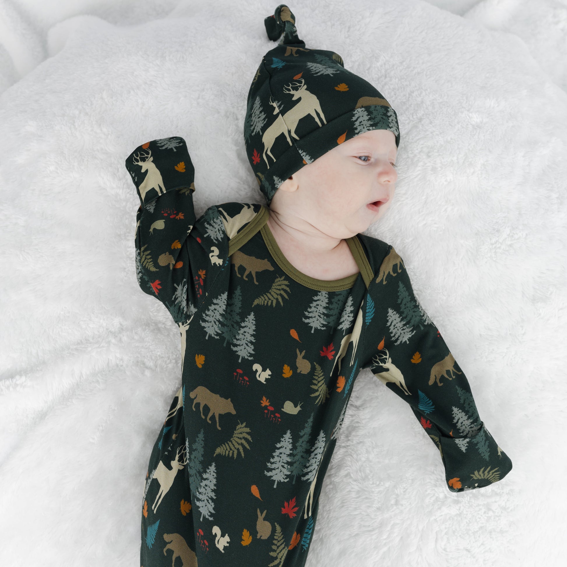 A baby lying on a white blanket, wearing a dark forest-print gown sleeper and matching beanie hat with woodland animal motifs.