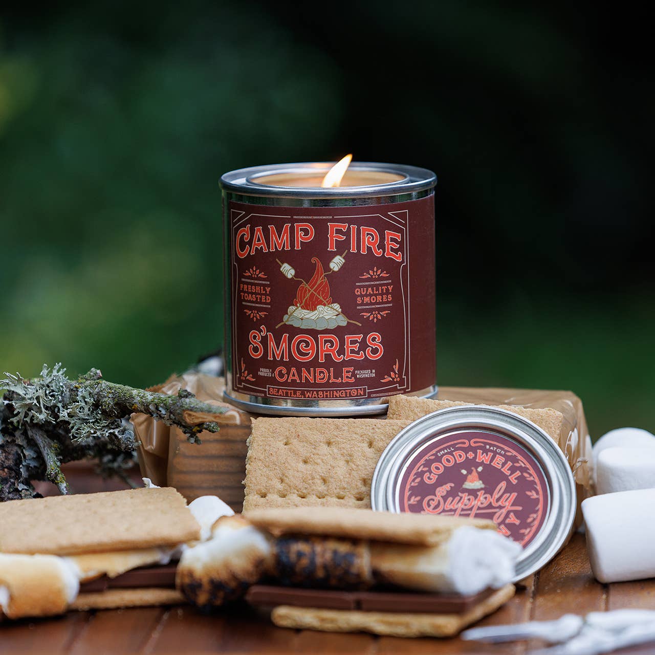 Campfire S'mores candle with graham crackers, marshmallows, and a stick on a wooden surface with a blurred green background.
