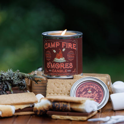 Campfire S'mores candle with graham crackers, marshmallows, and a stick on a wooden surface with a blurred green background.