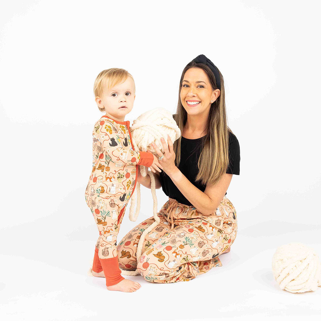 Mom and baby holding white ball of yarn while modeling our pet cat print that is orange with red/orange trim. Features cats, plants, books and yarn on the bamboo pajamas as well.