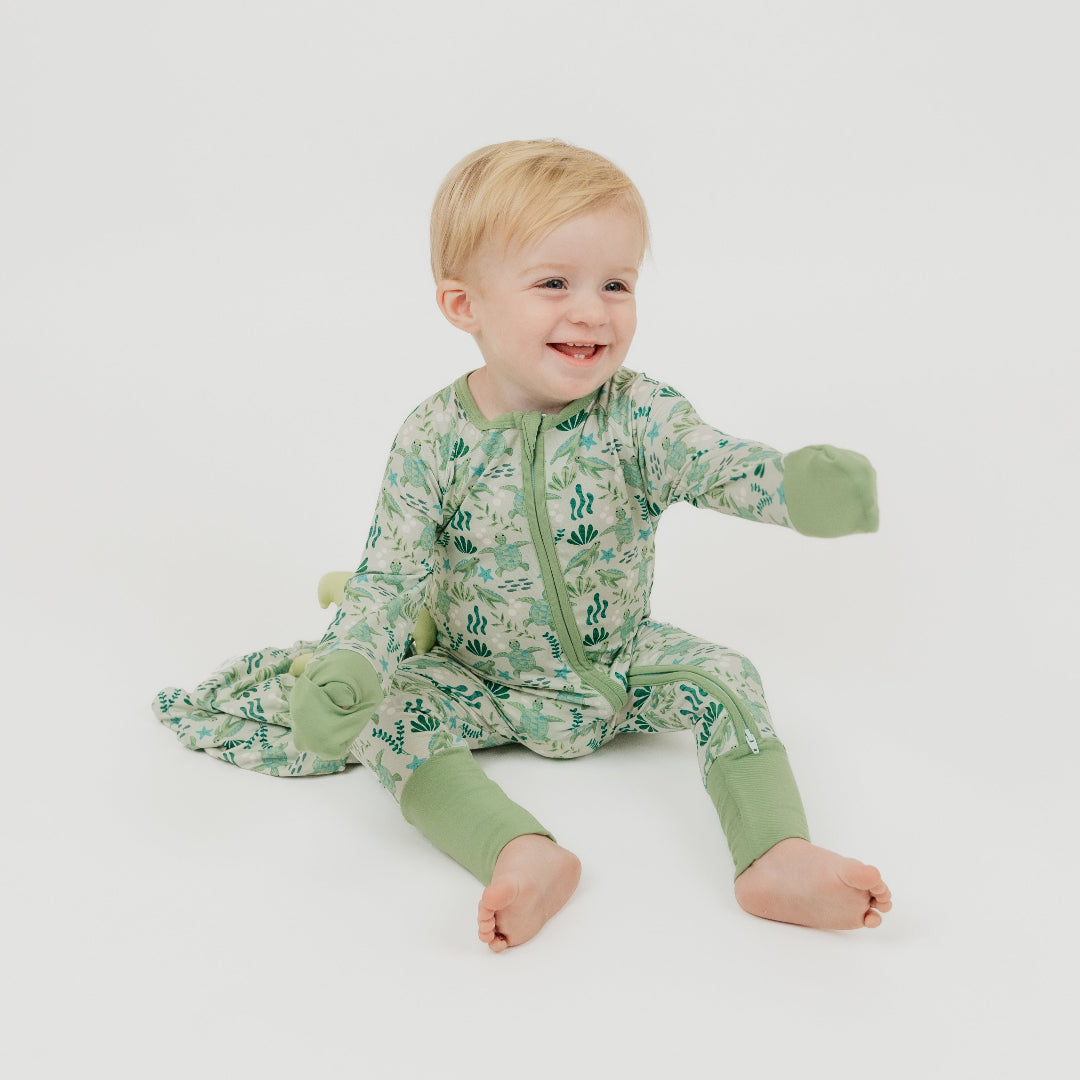 Baby boy sitting on a white background wearing our Save the Sea Turtles bamboo convertible baby pajamas by Emerson and Friends. This ultra-soft, eco-friendly footie features a charming sea turtle and ocean-inspired print. Made from breathable bamboo fabric, it’s perfect for sensitive skin and designed for ultimate comfort with fold-over feet and mittens.