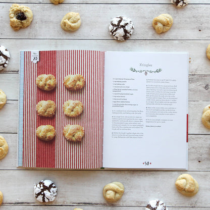 Cookbook open to a page with cookie recipes surrounded by various cookies on a wooden surface